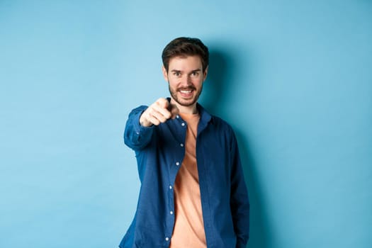 Confident smiling guy choosing or inviting you, pointing finger at camera decisive, standing on blue background.