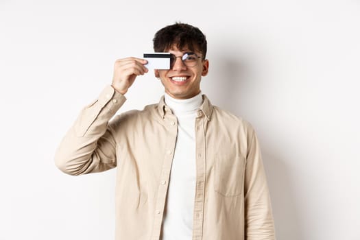 Cheerful young guy smiling real and showing plastic credit card, standing happy on white background, wearing glasses.