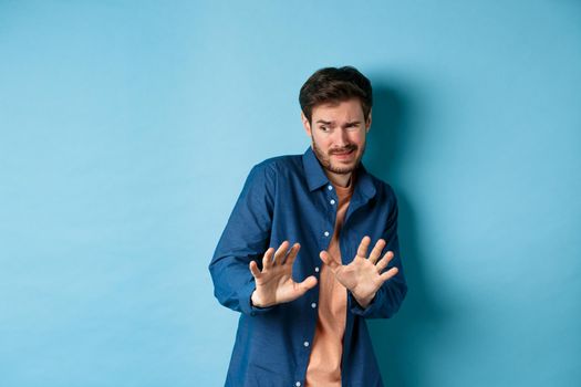 Man cringe and stay away from something nasty, looking aside and stretch out hands to reject bad deal, standing on blue background.