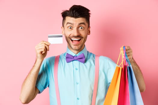 Excited man showing plastic credit card and shopping bags, smiling amazed while buying gifts, standing on pink background.