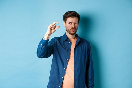 Image of young caucasian man showing small size and squinting to see something tiny, standing on blue background.