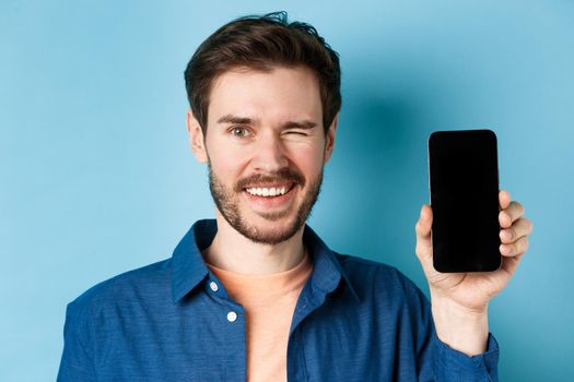 Close-up of handsome young man smiling, winking and showing empty mobile phone screen, standing in casual clothes on blue background.