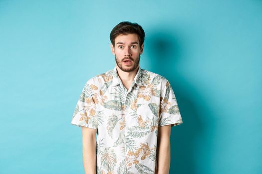 Image of shocked tourist guy drop jaw, gasping and looking startled, standing in hawaiian shirt on blue background. Copy space