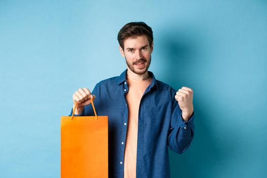 Handsome smiling man saying yes, triumphing and showing shopping bag, celebrate good promo offer, standing on blue background.
