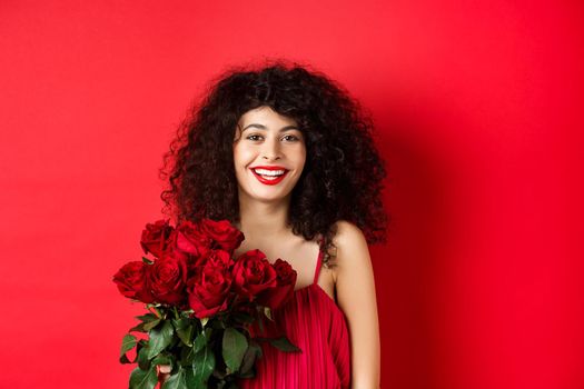 Happy fashionable female model with bouquet of red roses, smiling and looking cheerful at camera, studio background.