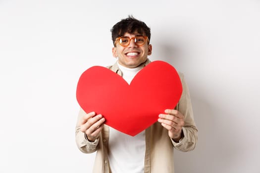 Cheerful young man smiling and looking thrilled at camera, showing big red heart cutout on Valentines day, making romantic gift to lover, white background.