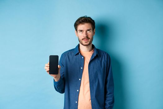 Annoyed young guy showing empty smartphone screen and roll eyes up bothered, standing on blue background.