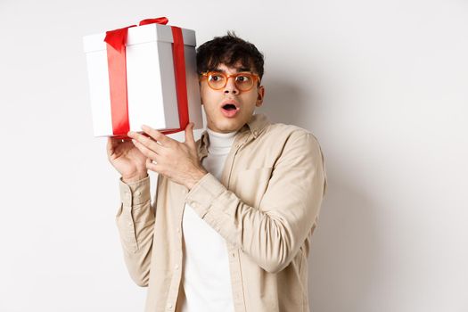 Holidays concept. Funny young man in glasses shaking gift box, listening what inside present and gasping amazed, standing on white background.