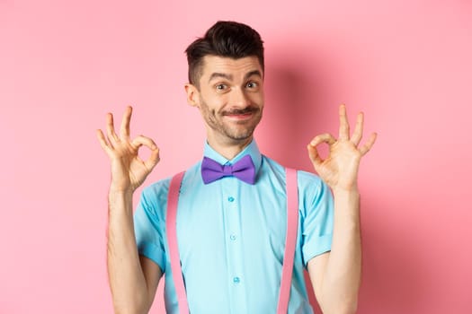 Portrait of handsome man with moustache and bow-tie, showing okay gestures and smiling in approval, recommending good offer, standing over pink background.