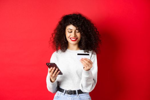 E-commerce and online shopping concept. Young modern woman paying with credit card, making purchase with smartphone, standing on red background.