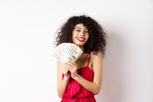 Excited woman in red dress winning money, showing dollar bills and smiling happy, standing on white background.