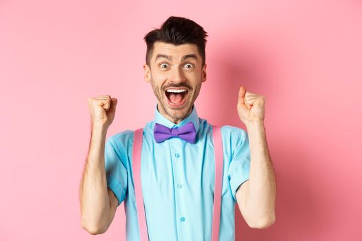 Happy guy winning prize, screaming with joy yes, shaking hands and celebrating victory, achieve success and triumphing over pink background.