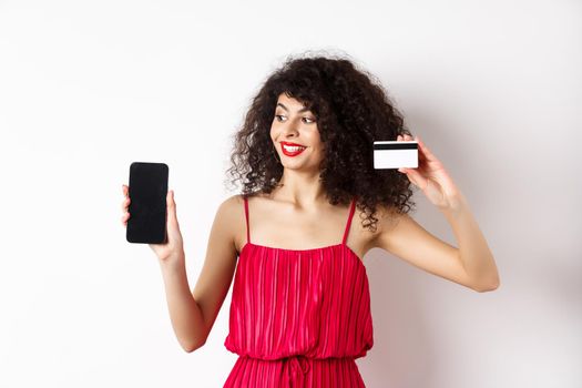 Online shopping concept. Elegant curly-haired woman in red dress showing plastic credit card and empty mobile phone screen, standing over white background.