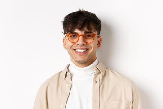 Close-up of handsome hipster guy with natural smile, wearing glasses and earrings, looking happy at camera, standing on white background.