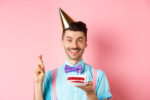 Holidays and celebration concept. Happy young man enjoying birthday party, wearing cone hat and cross fingers, making wish on bday cake with candle, pink background.