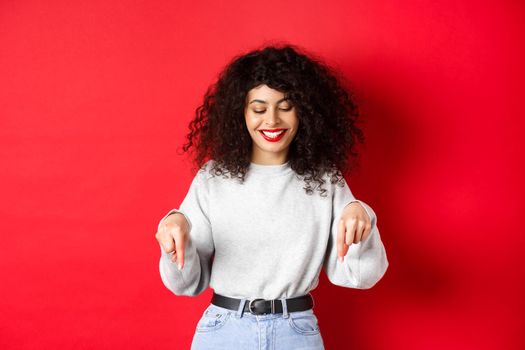 Modern woman with curly hairstyle and red lips, pointing fingers down and smiling happy, showing advertisement on empty space, studio background.