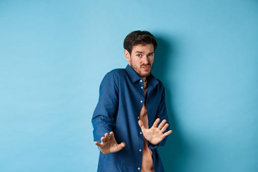 Timid young man asking to keep away, stretch out hands defensive and step back, refusing something disgusting, standing on blue background.
