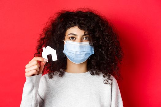Real estate and pandemic concept. Young woman in medical mask showing small paper house cutout, standing on red background.