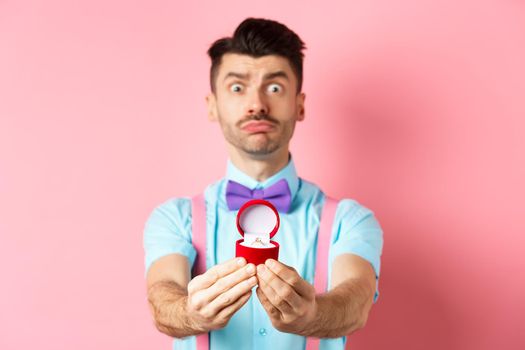 Valentines day. Nervous boyfriend waiting for girlfriend reply on marriage proposal, showing engagement ring and looking worried, standing over pink background.