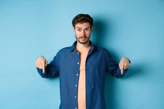 Confused male model in casual clothes, looking puzzled and pointing fingers down, showing banner, standing on blue background.
