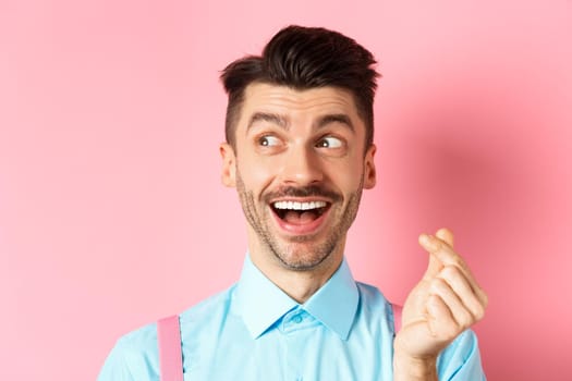 Valentines day concept. Romantic guy showing finger heart and looking left, smiling happy, standing on pink background dreamy.