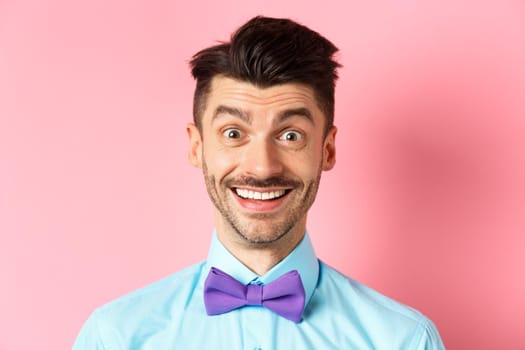 Close-up of cheerful caucasian man smiling happy, looking at something interesting, standing in bow-tie over pink background.