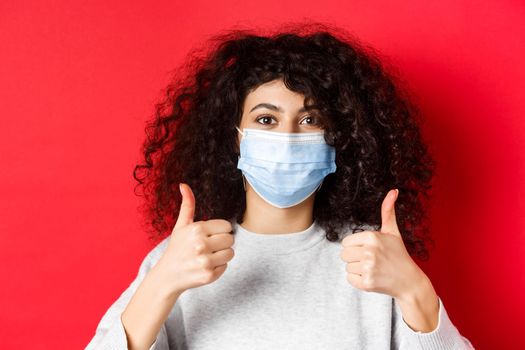 Covid-19 and pandemic concept. Close-up of young curly woman in face mask showing thumbs up, like and praise good thing, standing on red background.