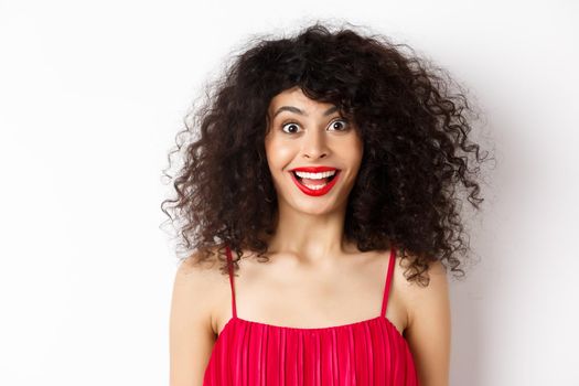 Close-up of happy lady with curly hair and red lips, raising eyebrows and looking surprised at camera, standing over white background.