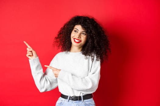 Beautiful european woman with curly hair and makeup, pointing fingers left and looking aside with dreamy smile, checking out promotion deal, red background.