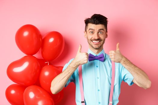 Valentines day concept. Smiling caucasian man showing thumbs up in approval, recommending special deal for lover, standing near cute red hearts balloons and pink background.