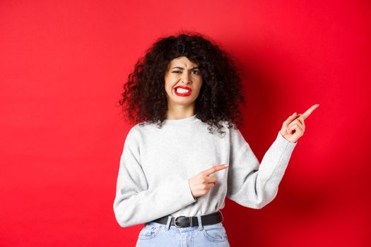 Skeptical caucasian woman grimacing dissasitsfied, complain on bad taste, pointing fingers right and cringe from dislike, standing against red background.