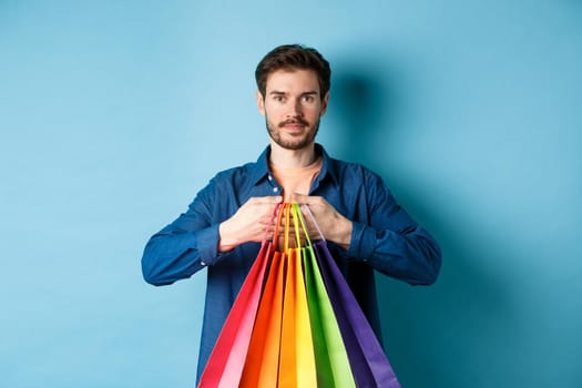 Modern guy with beard, holding packages from shop and smiling, standing on blue background.