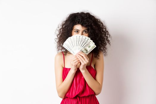 Gorgeous woman with curly hair, hiding face behind dollars and smiling, standing over white background. Copy space