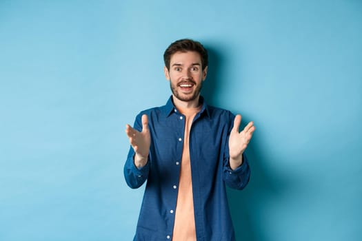 Cheerful young man pointing hands at camera and smiling, feeling glad for you, praising nice work, standing on blue background.