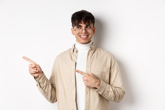 Handsome male model in glasses and trendy clothes, looking and pointing left at empty space, smiling with happy and pleased face expression, white background.