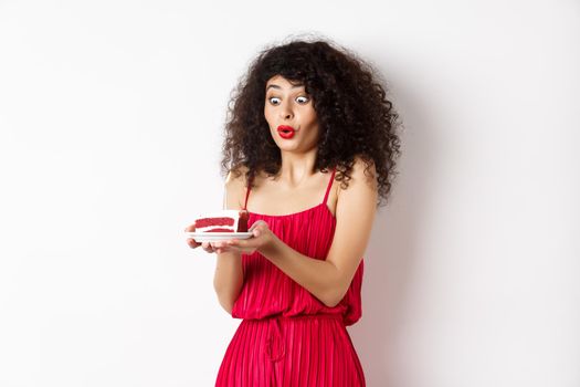 Excited birthday girl in red dress blowing candle on cake and making wish, standing on white background.