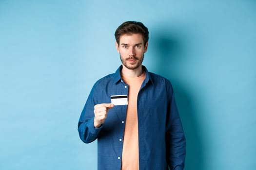 Handsome young man with beard, wearing casual clothes, showing plastic credit card and looking at camera, blue background.