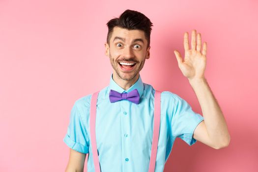 Friendly smiling man in funny bow-tie saying hello, waving hand to greet you, make hi gesture and looking happy yo see you, standing over pink background.