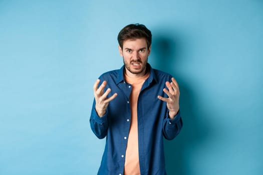 Annoyed and angry man raising hands and clench teeth outraged, stare pissed-off at camera, going to kill someone, standing on blue background.