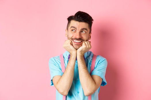Attractive smiling guy dreaming of something, looking at upper left corner imaging things, daydreaming on pink background. Copy space