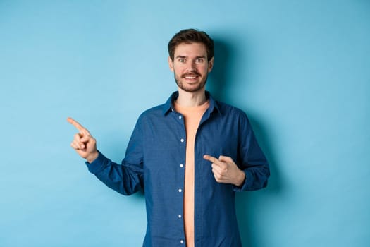 Happy smiling caucasian man showing advertisement, pointing fingers left at empty space, standing on blue background.