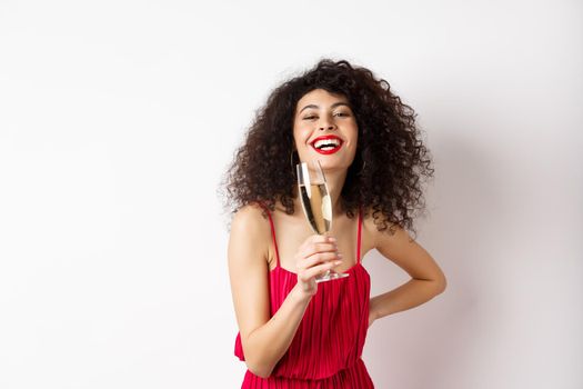 Cheerful elegant woman with dark curly hair, wearing party dress, celebrating anniversary on valentines day, drinking champagne from glass and smiling at camera, white background.