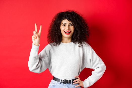 Young pretty lady in sweatshirt showing number two, making an order and smiling, standing on red background.