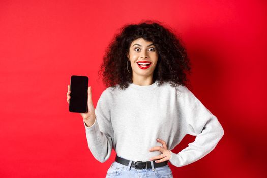 Excited woman with curly hair and red lips, showing empty smartphone screen and screaming from joy, standing on red background.