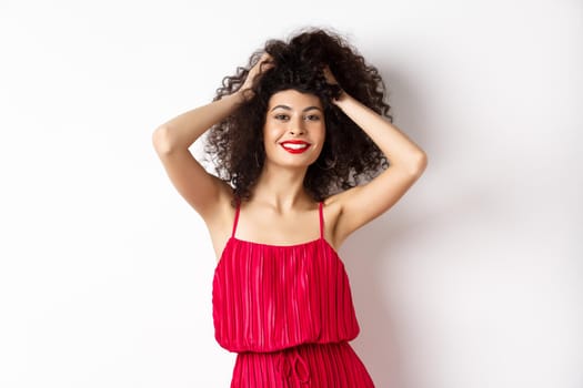 Beauty and fashion. Carefree woman in red dress and makeup, touching curly hair and smiling happy, standing on white background.