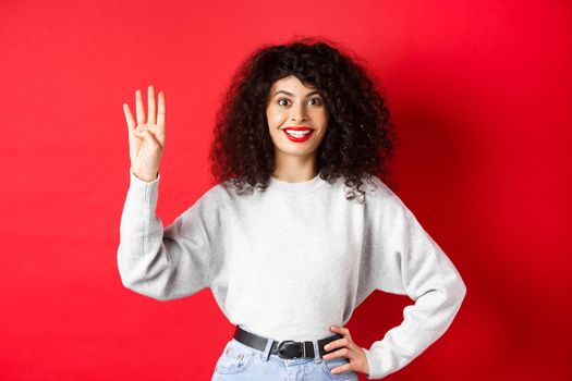 Excited woman with curly hair showing number four with fingers, making order, standing against red background.