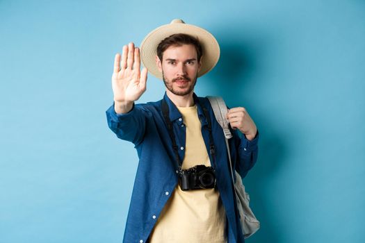 Serious male tourist warn you, stretch out hand to show stop gesture. Man on vacation forbid something, refusing or prohibiting action, standing with backpack on blue background.