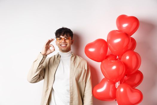 Valentines and romance concept. Happy young man smiling, showing OK sign near red hearts gesture, recommending good offer on lovers day.