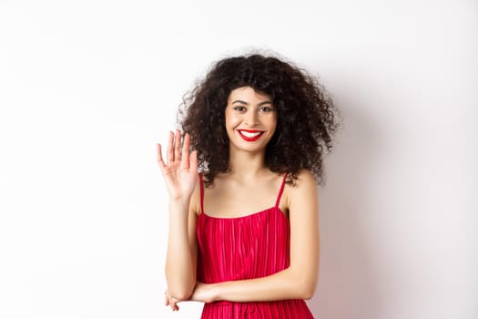 Cheerful elegant woman saying hello, waving hand and smiling at camera, greeting someone, standing in red dress on white background.