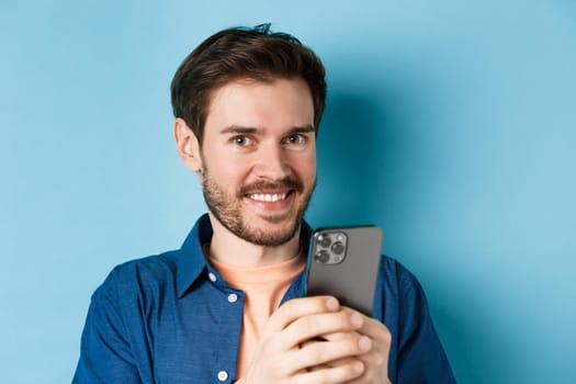Close up of young guy with beard, smiling happy at camera and holding mobile phone, standing on blue background.